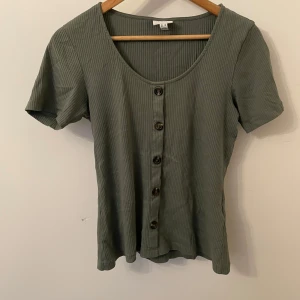 Grön ribbad topp från Aware by Vero Moda - Säljer en stilren grön ribbad topp från Aware by Vero Moda. Toppen har korta ärmar och knappar framtill, perfekt för en avslappnad look.