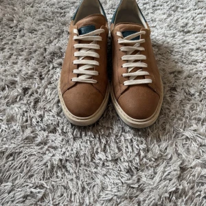 Brunello Cucinelli sneakers i brunt skinn - Snygga sneakers från Brunello Cucinelli i brunt skinn med vita snören och en kontrasterande blå detalj vid hälen. Perfekta för en stilren look. Skorna är gjorda i 100% kalvskinn vilket gör de mycket exklusiva och mycket bekväma på foten. Nypris ca 9000