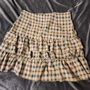 Rutig kjol med volanger - Charmig rutig kjol i beige och blå toner med volanger. Kjolen har en kort längd och är perfekt för en trendig look. Tillverkad i ett lätt och bekvämt material.