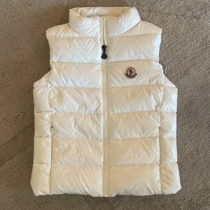 Vit dunväst från Moncler (barn strl 140/10 år)) - Säljer en stilren vit barndunväst från Moncler (Ghany Gilet) med dragkedja och hög krage. Perfekt för kyliga dagar när du vill hålla värmen utan att kompromissa med stilen. 