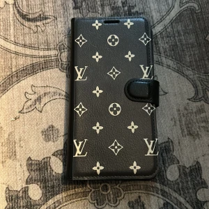 Nytt mobilskal! Svart plånboksväska med monogram, till iPhone 16 plus  - Snygg svart plånboksväska med ikoniskt monogrammönster. Väskan har plats för kort och en praktisk stängning med knapp. Perfekt för att skydda din telefon med stil. Mobilskal till iPhone 16 plus! Nytt! 