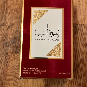 Ameerat Al Arab Eau de Parfum från Asdaaf - Elegant parfymflaska i röd sammet med gulddetaljer. Ameerat Al Arab är en Eau de Parfum med en rik och orientalisk doftprofil. Flaskan rymmer 100 ml och har en dekorativ guldfärgad kork med intrikata mönster.