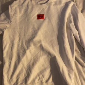 Vit sweatshirt från Hugo - Säljer en stilren vit sweatshirt från Hugo med röd logga på bröstet. Tröjan har långa ärmar och ribbade muddar vid ärmslut och nederkant. Perfekt för en avslappnad look. 