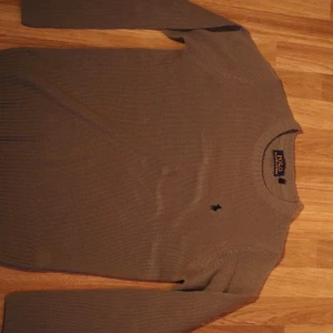 Grå ribbad tröja från Polo Ralph Lauren - Snygg grå ribbad tröja från Polo Ralph Lauren med långärmad design. Perfekt för en stilren och avslappnad look. Den har en diskret logga på bröstet.