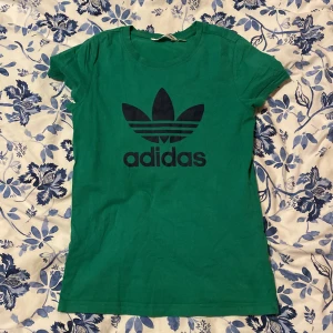 Grön t-shirt från Adidas - Säljer en grön t-shirt från Adidas med det klassiska logotyptrycket i svart på framsidan. T-shirten är kortärmad och tillverkad i bomull, perfekt för en avslappnad stil.