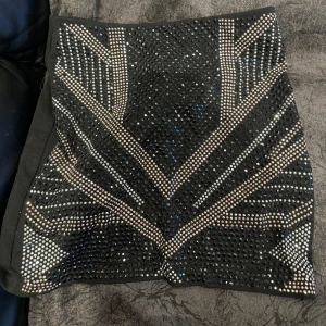 Svart glittrig kjol från H&M - Snygg svart kjol från H&M med glittriga detaljer i ett geometriskt mönster. Perfekt för att sticka ut med stil. Kjolen har en tight passform och är kort i modellen. Helt oanvänd  med prislappen kvar nypris 299kr