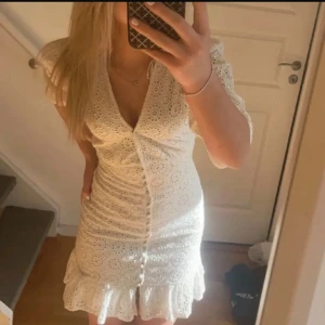 Vit klänning med spetsdetaljer - Charmig vit klänning med spetsdetaljer och volangkant. Den har en v-ringad design och korta ärmar. Perfekt för sommarens festligheter.