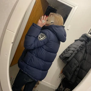 Blå d rand jacka  - Säljer en blå d brand med huva och ett märke på ärmen. Jackan har dragkedja och är perfekt för kyliga dagar. Den är vadderad och har en ljus insida för extra komfort. Storlek L men känns som M.