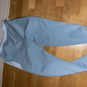 Ljusblå mjukisbyxor från Nike - Säljer ett par ljusblå mjukisbyxor från Nike med snörning i midjan och svarta dragkedjor på sidorna. Perfekta för en avslappnad stil.