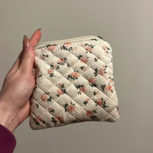 Blommig quiltad plånbok - Söt liten plånbok med blommigt mönster i rosa och grönt på en beige bakgrund. Den är quiltad och har en praktisk dragkedja upptill. Perfekt för att hålla ordning på småsaker i väskan.