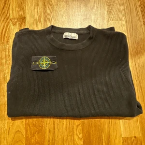 Svart tröja från Stone Island - Säljer en stilren svart tröja från Stone Island med ribbad struktur och deras ikoniska märke på ärmen. Perfekt för en casual look.