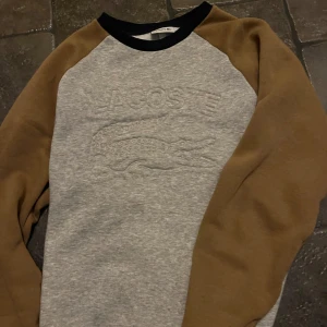 Grå och brun tröja från Lacoste - Säljer en snygg grå tröja från Lacoste med bruna ärmar och svart krage. Tröjan har ett diskret Lacoste-märke på bröstet och är perfekt för en avslappnad stil.