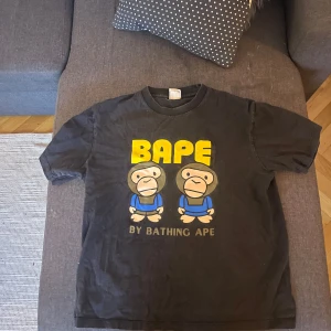 Svart t-shirt från BAPE - Svart t-shirt från BAPE med färgglatt tryck av två apor och texten 'BAPE BY BATHING APE' på både fram- och baksidan. Perfekt för streetwear-entusiaster. Det är en L men är liten i storleken så passar även S och M