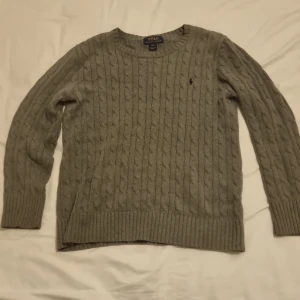 Grå kabelstickad tröja från Polo Ralph Lauren - Säljer en stilren grå kabelstickad tröja från Polo Ralph Lauren. Tröjan har långa ärmar och en klassisk rund halsringning. Perfekt för kyligare dagar och ger en tidlös look. Storlek l men passar 160 till 175 cm