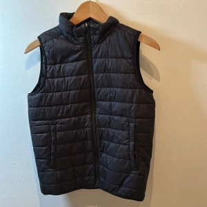 Svart dunväst från Everest - Säljer en svart dunväst från Everest med dragkedja framtill. Västen har en quiltad design och är perfekt för kyligare dagar. Den är ärmlös och har en hög krage för extra värme. Passar bra för både vardag och utomhusaktiviteter.