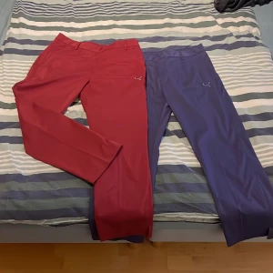 Röda och blå byxor från Puma - Två par byxor från Puma i rött och blått. De har en rak passform och är tillverkade i ett syntetmaterial. Byxorna har en diskret Puma-logga på sidan och är perfekta för en sportig look. Buda på, går att köp styckvis.