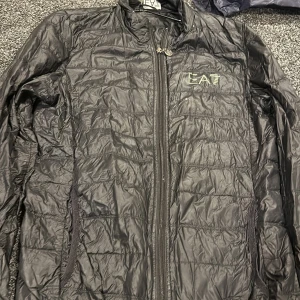Svart quiltad jacka från EA7 - Snygg svart quiltad jacka från EA7 med dragkedja framtill. Perfekt för kyligare dagar med sin lätta och stilrena design. Jackan har långa ärmar och en diskret logga på bröstet.