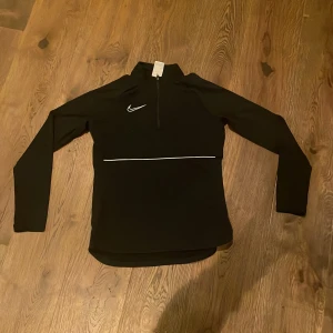 Svart långärmad tröja från Nike - Säljer en svart långärmad tröja från Nike. Tröjan har en dragkedja framtill och ett broderat Nike-märke på bröstet. Perfekt för träning och sportiga aktiviteter.