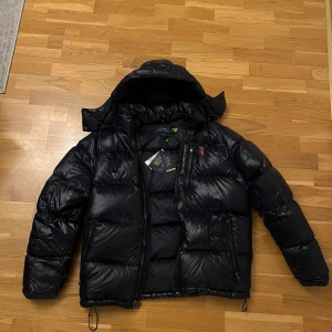 Navy blå dunjacka från Polo Ralph Lauren - Navy blå dunjacka från Polo Ralph Lauren med dragkedja och röd broderad logga på bröstet. Jackan har en pufferdesign och är perfekt för kalla dagar. Jackan är helt ny och inte använd, varför jag vill sälja den är för att den är för liten. Kan tänka mig sänka pris vid snabb affär 
