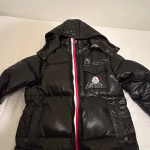 Svart dunjacka från Moncler - Snygg svart dunjacka från Moncler med huva och en iögonfallande röd och vit dragkedja. Jackan har en quiltad design och en praktisk bröstficka med Moncler-logga. Perfekt för kyliga dagar.