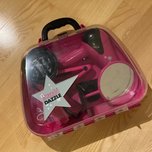 Leksak frisörs/styling-väska - Säljer ett rosa & svart frisörset i en praktisk väska. Setet innehåller en leksaksfön, spegel, 2 borstar, platttång och ett par hårspännen samt tofsar. Det är en del små rispor på väskan samt på en del av leksakerna i och den batteridrivna funktionen på hårfönen fungerar ej. I övrigt går allt bra att använda.