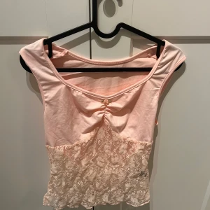 Rosa spetsdetaljerad topp -  rosa topp med spetsdetaljer och en liten rosett framtill. Toppen har en v-ringad design och är ärmlös.