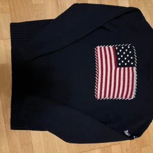 Mörkblå stickad tröja med amerikansk flagga - Säljer en mörkblå stickad tröja från north sails med en stor amerikansk flagga på framsidan. Tröjan har långa ärmar och ribbstickade muddar. Perfekt för en stilren och avslappnad look.
