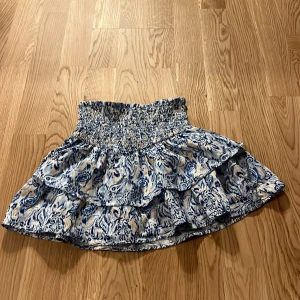 Blåmönstrad kjol från Gina Tricot i storlek 134-140 - Säljer en söt blåmönstrad kjol från Gina Tricot med volanger och elastisk midja. Perfekt för en somrig look med sitt livfulla mönster i blått och vitt.