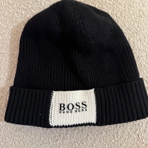 Svart mössa från Hugo Boss - Snygg svart mössa från Hugo Boss med ribbad design och vit etikett med logotyp framtill. Perfekt för att hålla värmen under kyliga dagar.