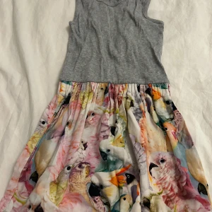 Grå ärmlös klänning med fågelmönster från Molo - Säljer en söt grå ärmlös klänning från Molo med ett färgglatt fågelmönster på kjolen. Perfekt för små äventyrare som älskar djur och färg. Klänningen har en ribbad överdel och en luftig kjol. Klänningen kommer tvättas och strykas innan den skickas! ⭐️ 