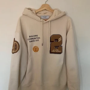 Beige hoodie från Les Deux - Snygg beige hoodie från Les Deux med tryck och broderade detaljer. Den har en stor siffra '2' och texten 'Building Communities Since 2011' på framsidan. Perfekt för en avslappnad stil.