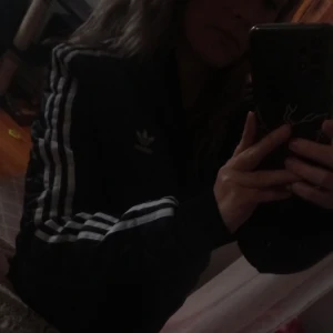 Svart quiltad bomberjacka från Adidas - Snygg svart quiltad bomberjacka från Adidas med de klassiska tre ränderna längs ärmarna. Jackan har dragkedja framtill och en sportig look. Perfekt för en casual stil.