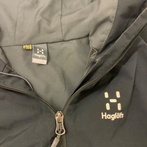 Haglöfsjacka - Säljer en svart vindjacka från Haglöfs med dragkedja framtill. Jackan har en enkel design med långärmade ärmar och är perfekt för blåsiga dagar. Den är funktionell och stilren, idealisk för utomhusaktiviteter. Storlek L