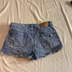 Blå jeansshorts från H&M Divided - Säljer ett par blå jeansshorts från H&M Divided i storlek 40. Shortsen har en cool slitning och knappgylf framtill. Perfekta för en avslappnad sommarlook!