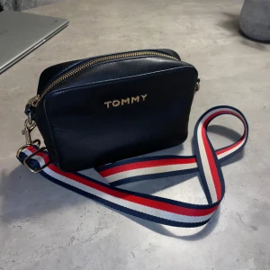 Tommy Hilfiger handväska - Snygg svart handväska från Tommy Hilfiger med ett justerbart axelband. Väskan har en dragkedja upptill och har fungerar perfekt för mig i att bära med nödvändigheter. Väskan har varit väl omhändertagen men har några tecken på användning (syns i kanterna på sista bilden) men som givetvis inte påverkar funktionen. Annars fräsch och fortfarande otroligt fin. 