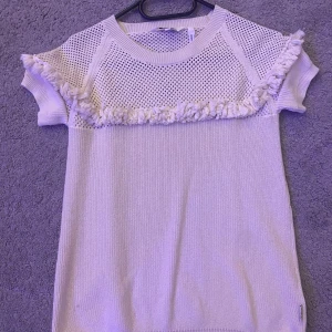 Vit stickad topp med fransdetaljer - Säljer en söt vit stickad topp med korta ärmar och fransdetaljer över bröstet. Toppen har ett nätliknande mönster upptill som ger en unik look. Perfekt för en avslappnad stil.