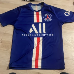 Blå PSG fotbollströja med Mbappe 7 - Säljer en blå PSG fotbollströja från Nike med Mbappe och nummer 7 på ryggen. Tröjan har korta ärmar och ett vitt och rött vertikalt mönster på framsidan. Perfekt för fans av Paris Saint-Germain!