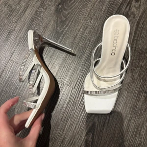 Vita klackskor från Boohoo - Eleganta vita klackskor från Boohoo med smala remmar dekorerade med glittriga detaljer. Skorna har en fyrkantig tå och en hög klack i silverfärg. Perfekta för att ge en stilfull touch till din outfit.
