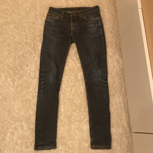 Mörkblå jeans från Nudie Jeans - Snygga mörkblå skinny jeans från Nudie Jeans med klassisk femficksdesign och orange sömmar. Perfekta för en stilren look. Midwaist och tillverkade i slitstarkt denim.