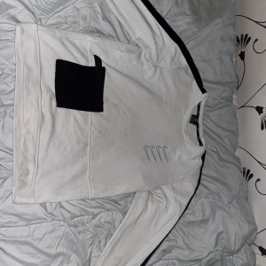 Vit långärmad tröja med svart tryck - Säljer en vit långärmad tröja med svart tryck och detaljer. Tröjan har en svart ficka på framsidan och svarta ränder längs ärmarna. Perfekt för en avslappnad stil.
