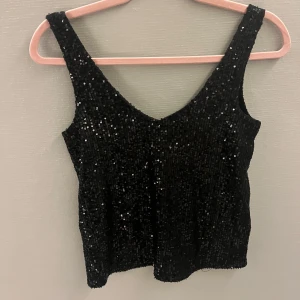 Svart glittrig topp från ONLY - Snygg svart glittrig topp från ONLY i storlek S. Perfekt för festliga tillfällen med sin glittriga yta och ärmlösa design. Toppen har en v-ringad hals och en tight passform. Perfekt till nyår!