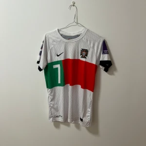 Portugal fotbollströja ”Ronaldo” - Snygg vit fotbollströja från Nike med nummer 7 och texten 'Ronaldo' på ryggen. Tröjan har korta ärmar och ett färgglatt mönster i grönt och rött på framsidan. Perfekt för fotbollsfans!