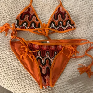 Färgglad bikini från Duga Adore - Snygg och färgglad bikini från Duga Adore med vågigt mönster i orange, rött och svart. Bikinin har knytband på sidorna och vid nacken för justerbar passform. Perfekt för sommarens stranddagar! aldrig använd 