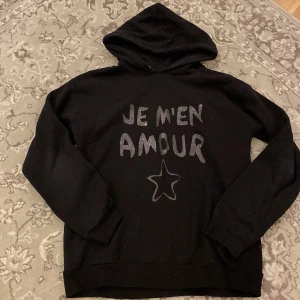 Svart hoodie från Gina Tricot - Säljer en svart hoodie från Gina Tricot med texten 'Je m'en amour' och en stjärna i glittrigt tryck på framsidan. Perfekt för en avslappnad stil med en touch av glitter. 🖤✨