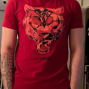 Röd t-shirt med tigertryck från Plein Sport - Snygg röd t-shirt från Plein Sport med ett coolt tigertryck i svart och orange på bröstet. Perfekt för dig som vill sticka ut med en djärv stil. Kortärmad och i en bekväm passform.