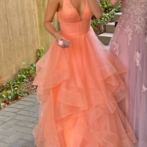 Persikofärgad balklänning med volanger - Elegant persikofärgad balklänning med djup v-ringning och flera lager av volanger. Klänningen har en tight passform upptill och en lång, flödande kjol. Perfekt för speciella tillfällen.