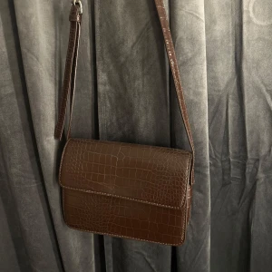 Brun axelväska med krokodilmönster - Snygg brun axelväska från Pieces med krokodilmönster. Väskan har en justerbar axelrem och stängs med en tryckknapp. Perfekt för att ge en stilren touch till din outfit.