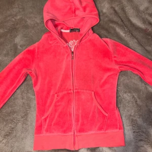 Röd velour hoodie från Juicy Couture - Säljer en snygg röd velour hoodie från Juicy Couture med dragkedja och huva. Den har ett stort, dekorativt emblem på ryggen i blått och vitt med glittriga detaljer. Perfekt för en avslappnad och trendig look. Den har några defekter och en fläck, som kanske går bort i tvätten! Massa stenar ramlat av också som inte synns!💕