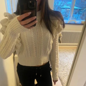 Vit stickad tröja med volangdetaljer - Säljer en snygg vit stickad tröja med volangdetaljer på ärmarna. Tröjan är croppad och har långa ärmar.