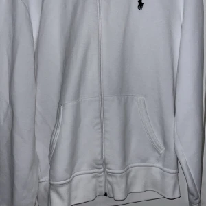 Vit hoodie från Ralph Lauren - Säljer en stilren vit hoodie från Ralph Lauren med dragkedja och det klassiska logotypen på bröstet. Perfekt för en avslappnad look.